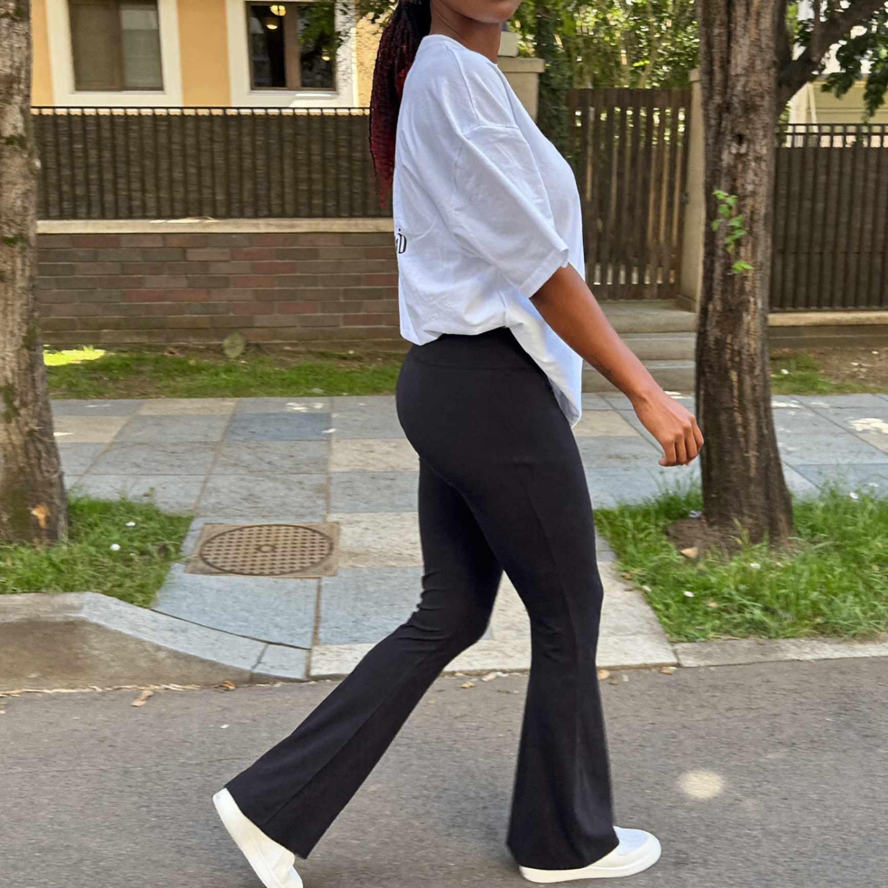 Cliente caminhando na rua usando a calça Noir preta flare com camiseta branca e tênis, mostrando o caimento de perfil.